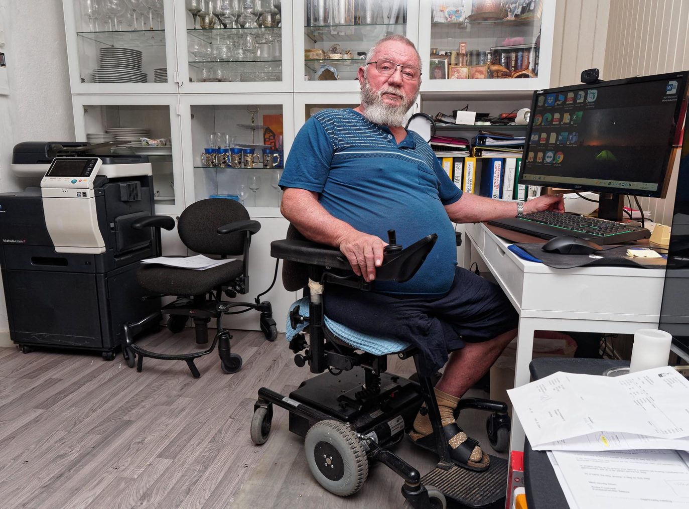 Nordby sitter i en elektrisk inne-rullestol ved et skrivebord med en pc i stuen.