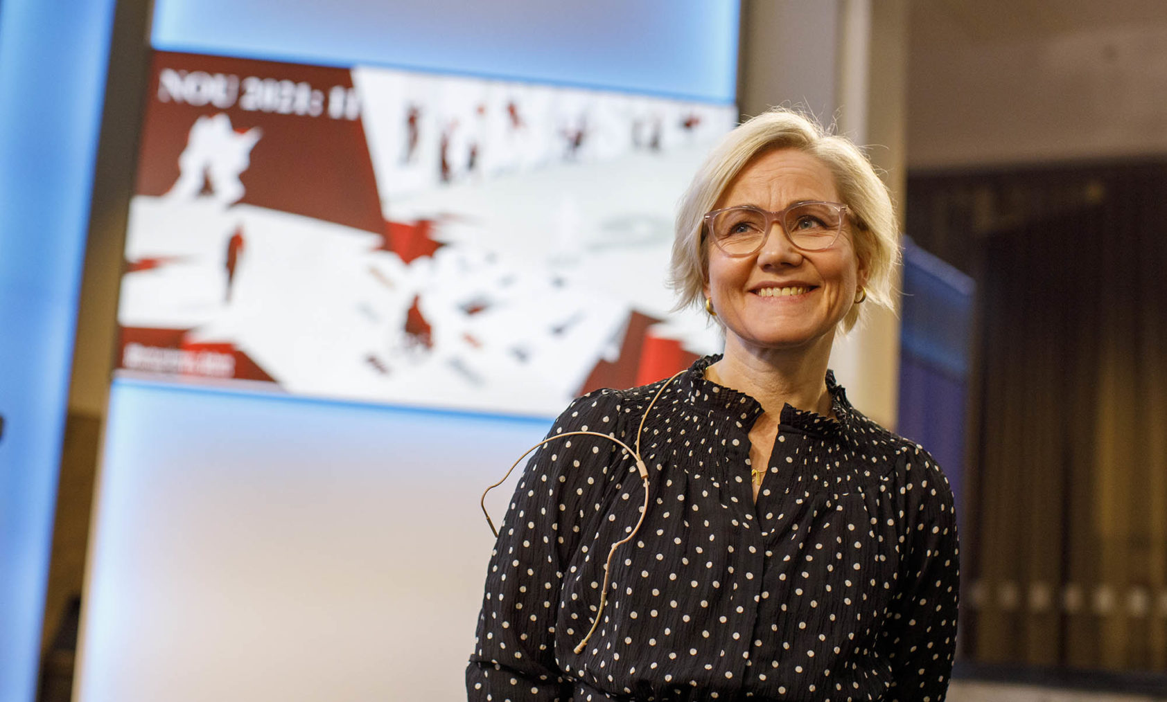 Ingvild Kjerkol med forsiden på utredningen uskarpt på en skjerm i bakgrunnen.