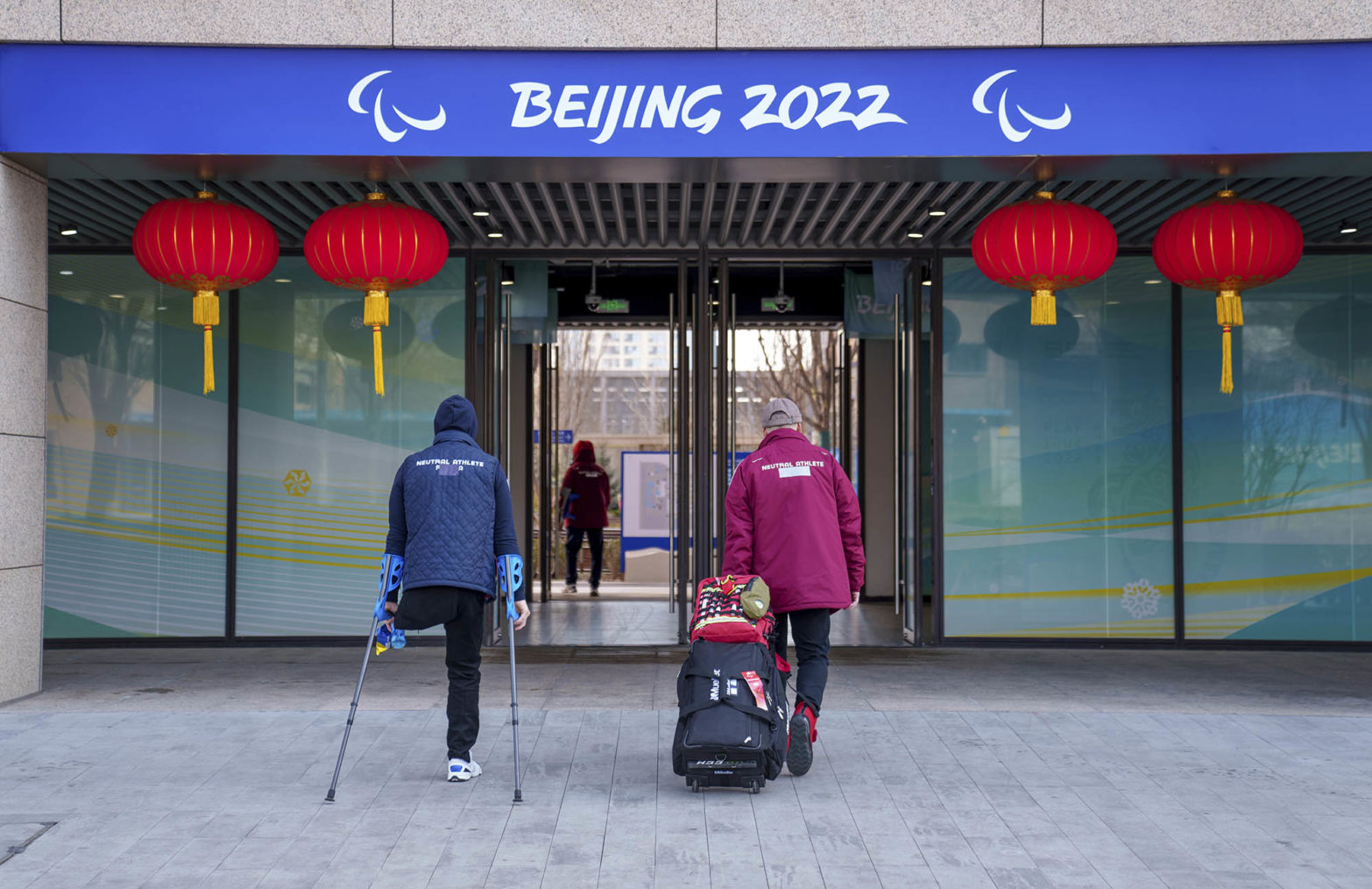 To peroner på vei inn i bygning med teksten «Beijing 2022» over døra, som er prydet med kinesiske lykter. Den ene mangler et bein og går på krykker.