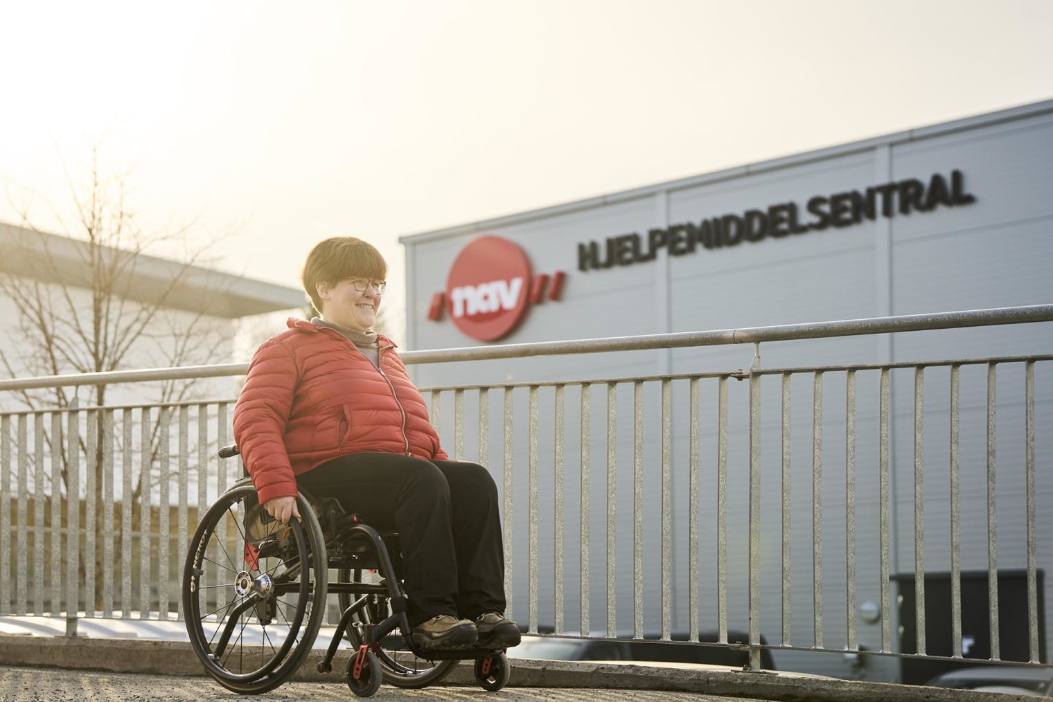 Ann-Helen Skogly ruller i rullestol forbi en bygning med skiltet Nav Hjelpemiddelsentral på veggen.