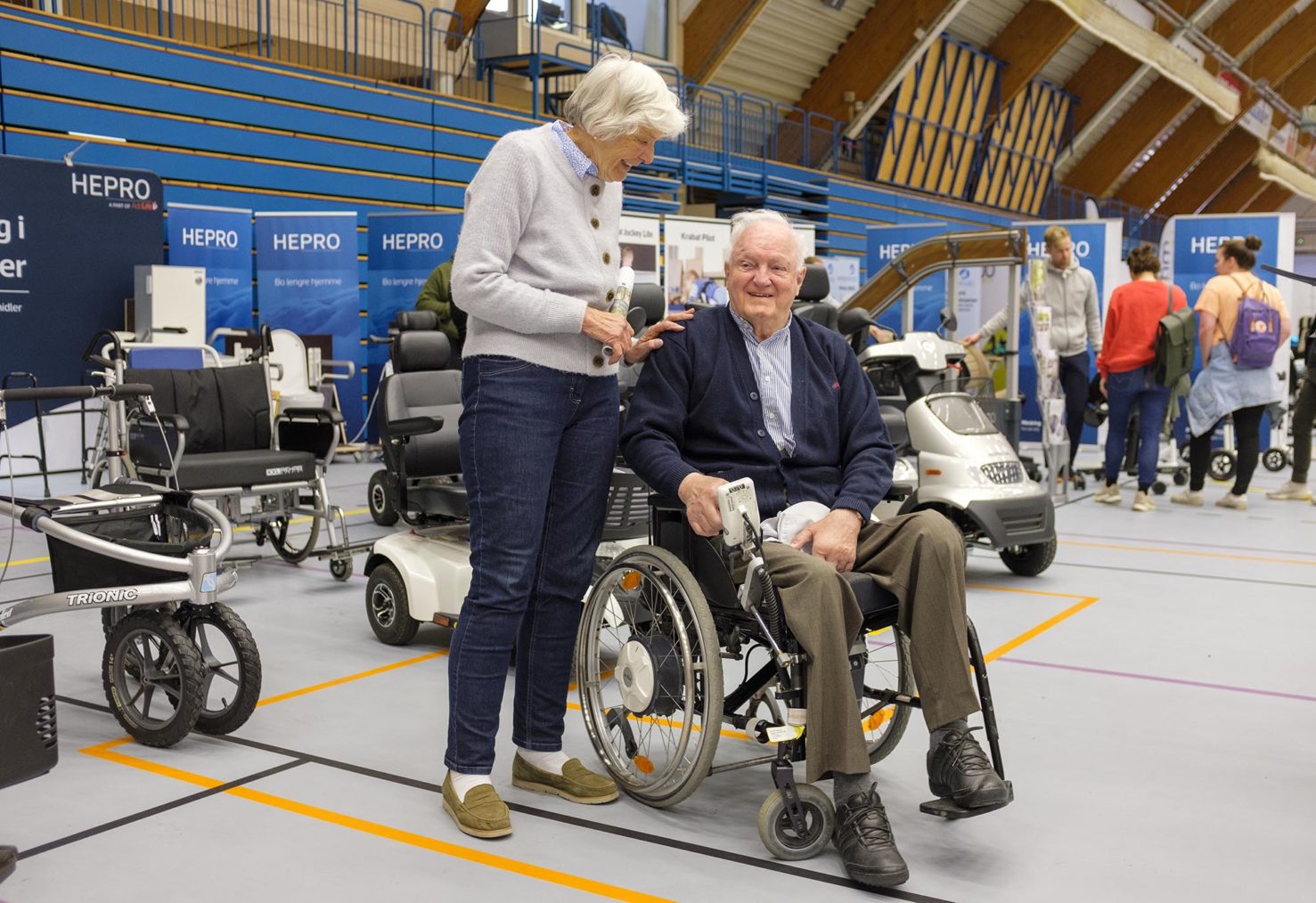 Ekteparet i samtale på stand med flere rullestoler. Han sitter i rullestol, hun står ved siden av.