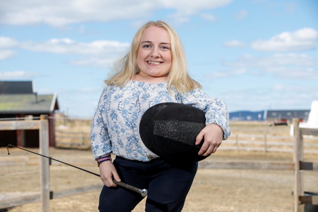 Hanna portrettert med ridehjelm og -pisk i hånden på en gård.