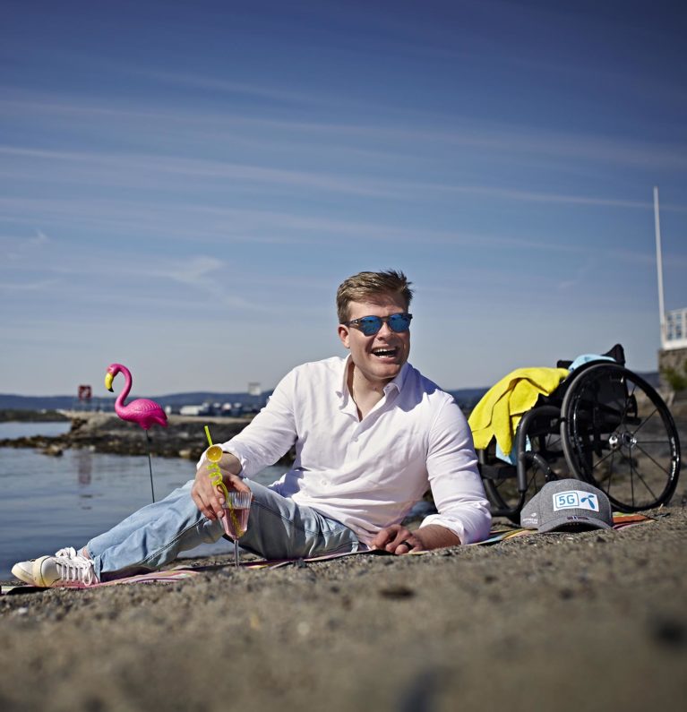 Jesper ligger på stranden og holder en drink. Han har solbriller på, og i bakgrunnen ser vi rullestolen og en rosa flamingo av plast.