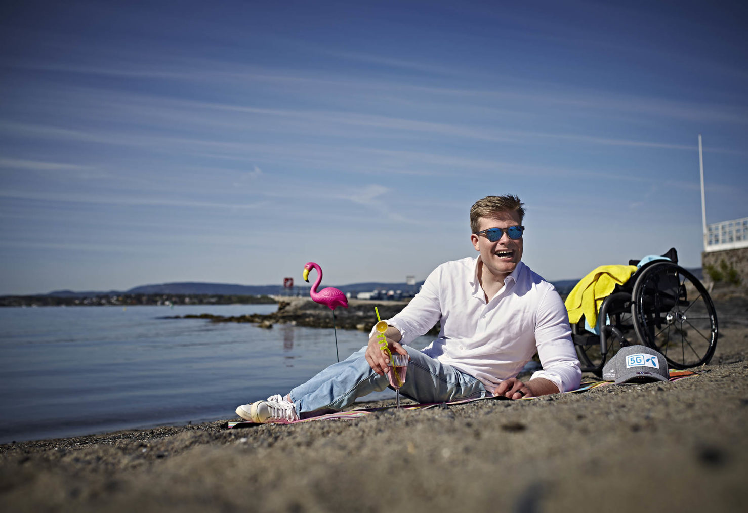 Jesper ligger på stranden og holder en drink. Han har solbriller på, og i bakgrunnen ser vi rullestolen og en rosa flamingo av plast.