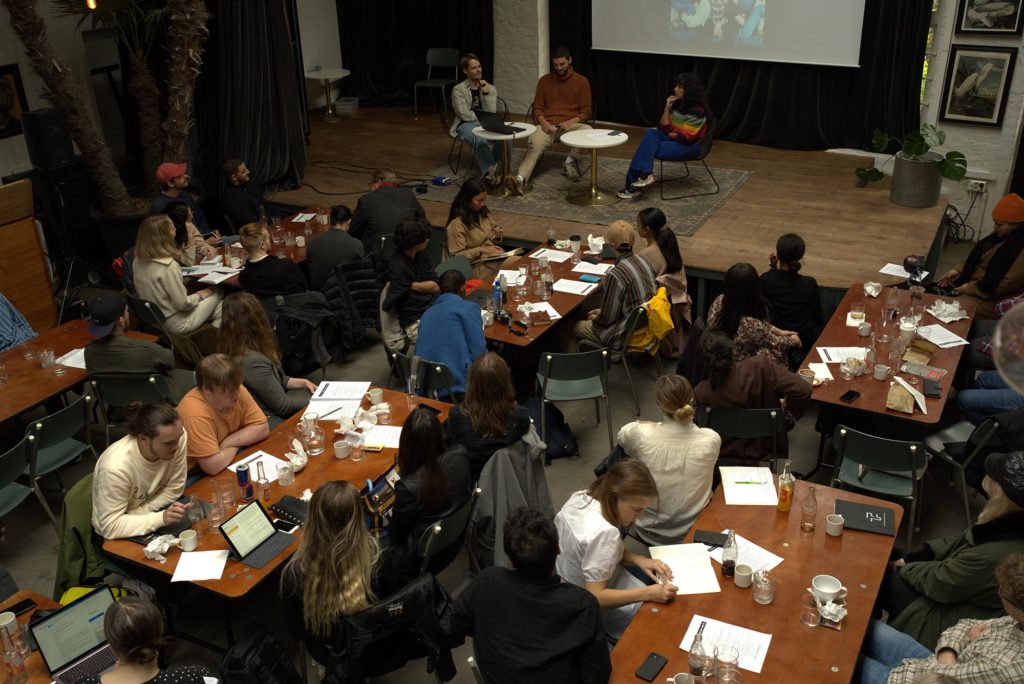 Bilde av en fullsatt møtesal der deltakerne sitter ved bord og panelet på et podium fremst.