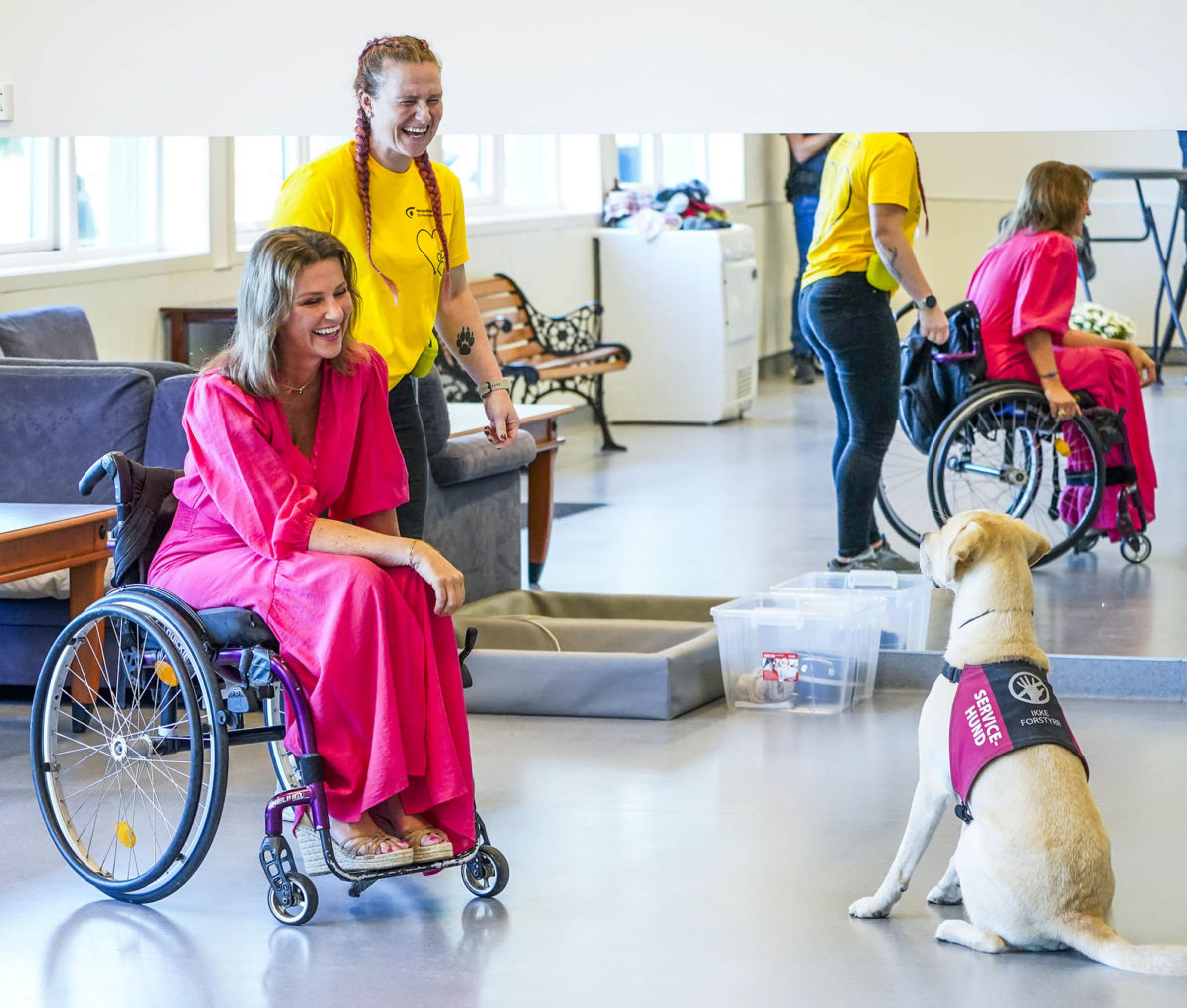 Märtha Louise sitter i en rullestol og smiler mot en hund som med "service dog"-skilt.