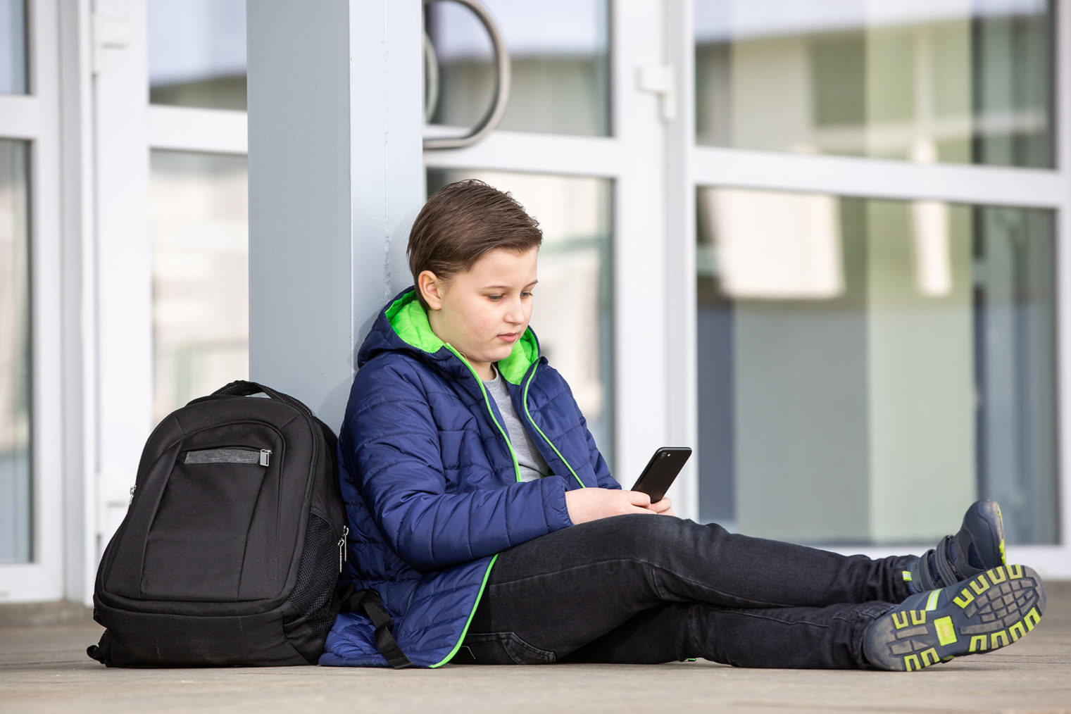 Skolegutt sitter utenfor skolebygningen og ser på telefonen sin. Sekken hans ligger på bakken ved siden av ham.