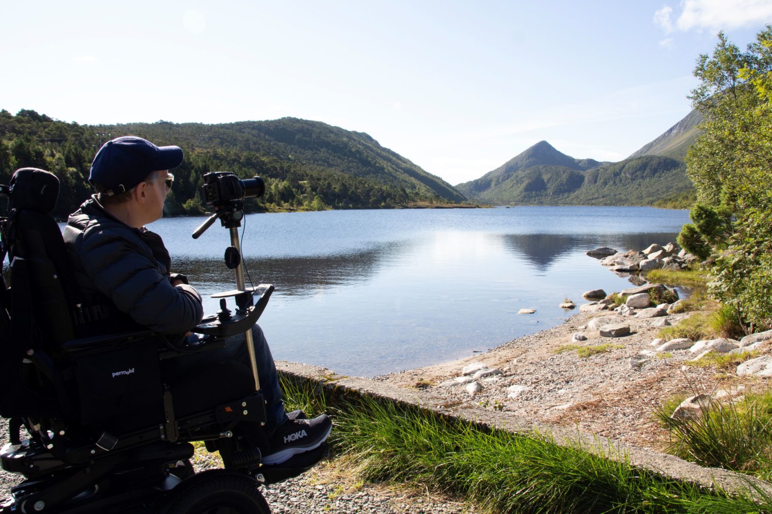 Richard Sundnes i rullestol foran vann og fjell rundt. Med kamera på stativ.