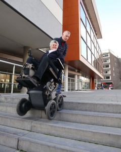 Eldre kvinne i rullestolen får bistand av en assistent til å rygge opp en trapp utendørs.