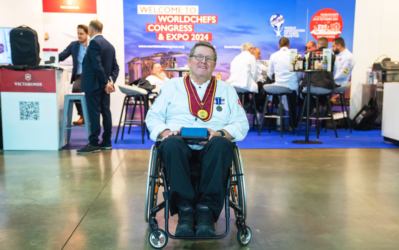 Tom Atle Steffensen poserer i rullestol med prisen foran en stand med World Chefs-plakat.
