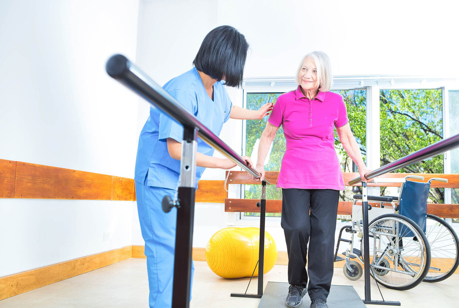 Eldre kvinne trener med fysioterapeut ved å gå i en skranke. I bakgrunnen står en rullestol.