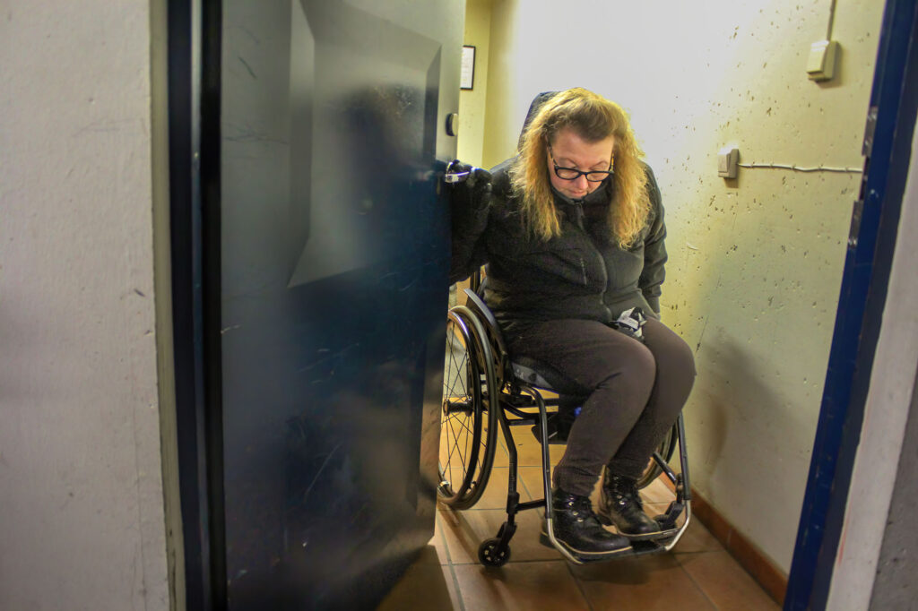 Pia i rullestol strever med å åpne døren og komme seg gjennom.