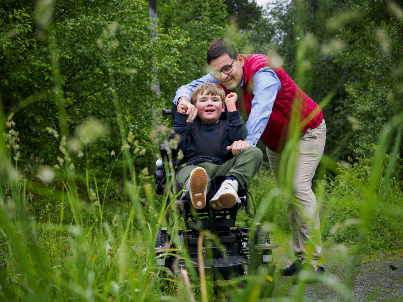 Far og sønn sammen i grønne omgivelser. Anders – i elektrisk rullestol – ler mot kamera. Håvard bøyer seg mot ham og smiler.