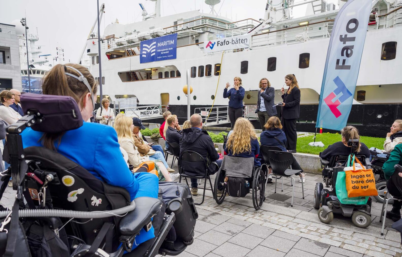 Bilde av møtet med rullestolbrukere i forgrunnen og båten i bakgrunnen.