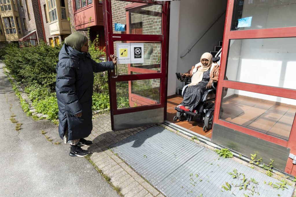 På vei ut inngangsdøren med rullestolen. Assistenen holder døren.