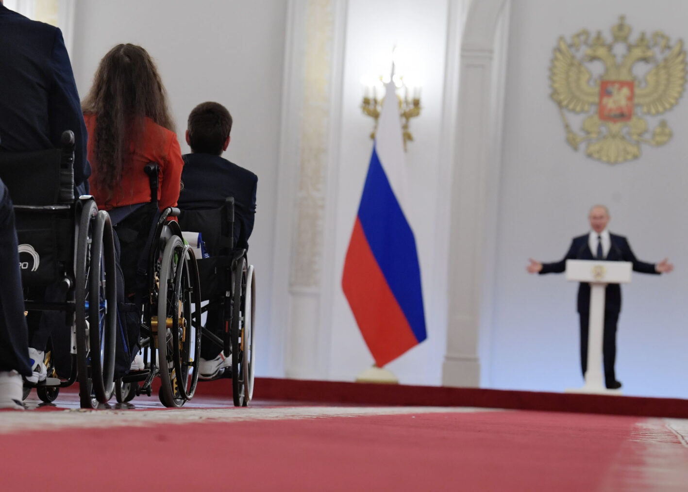 Gruppe av rullestolbrukere i forgrunnen. Putin på talerstol i bakgrunnen, ved siden av russisk flagg og riksvåpen.