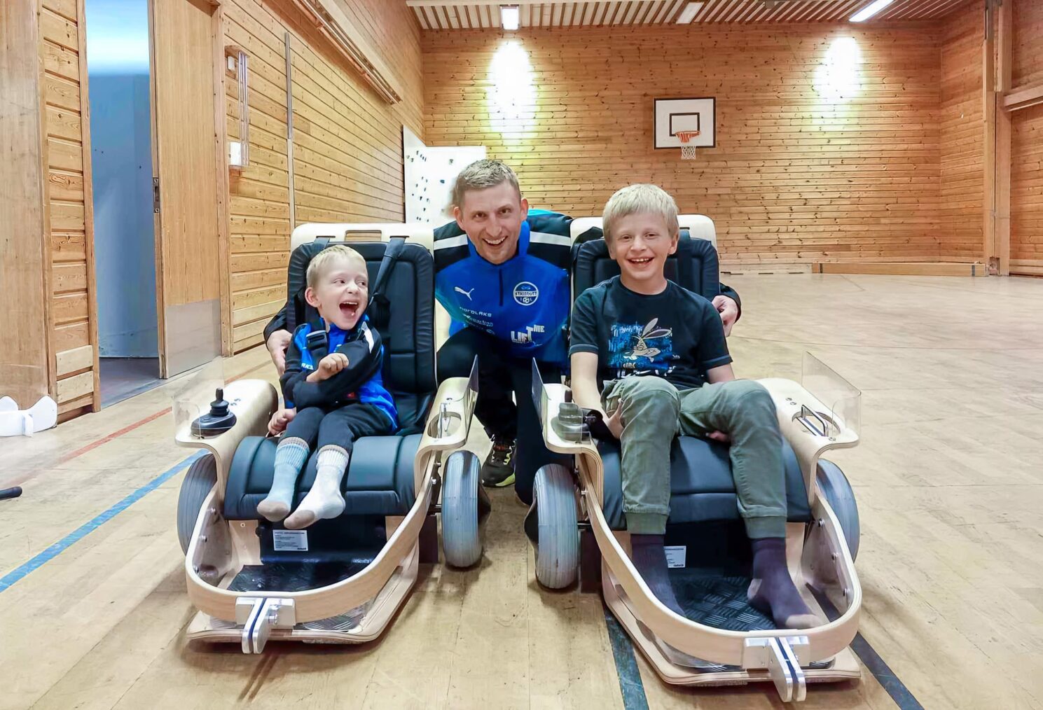 Gutta i el-innebandy-kjelker i en idrettshall. Treneren på huk bak. Alle smiler og ser i kamera.