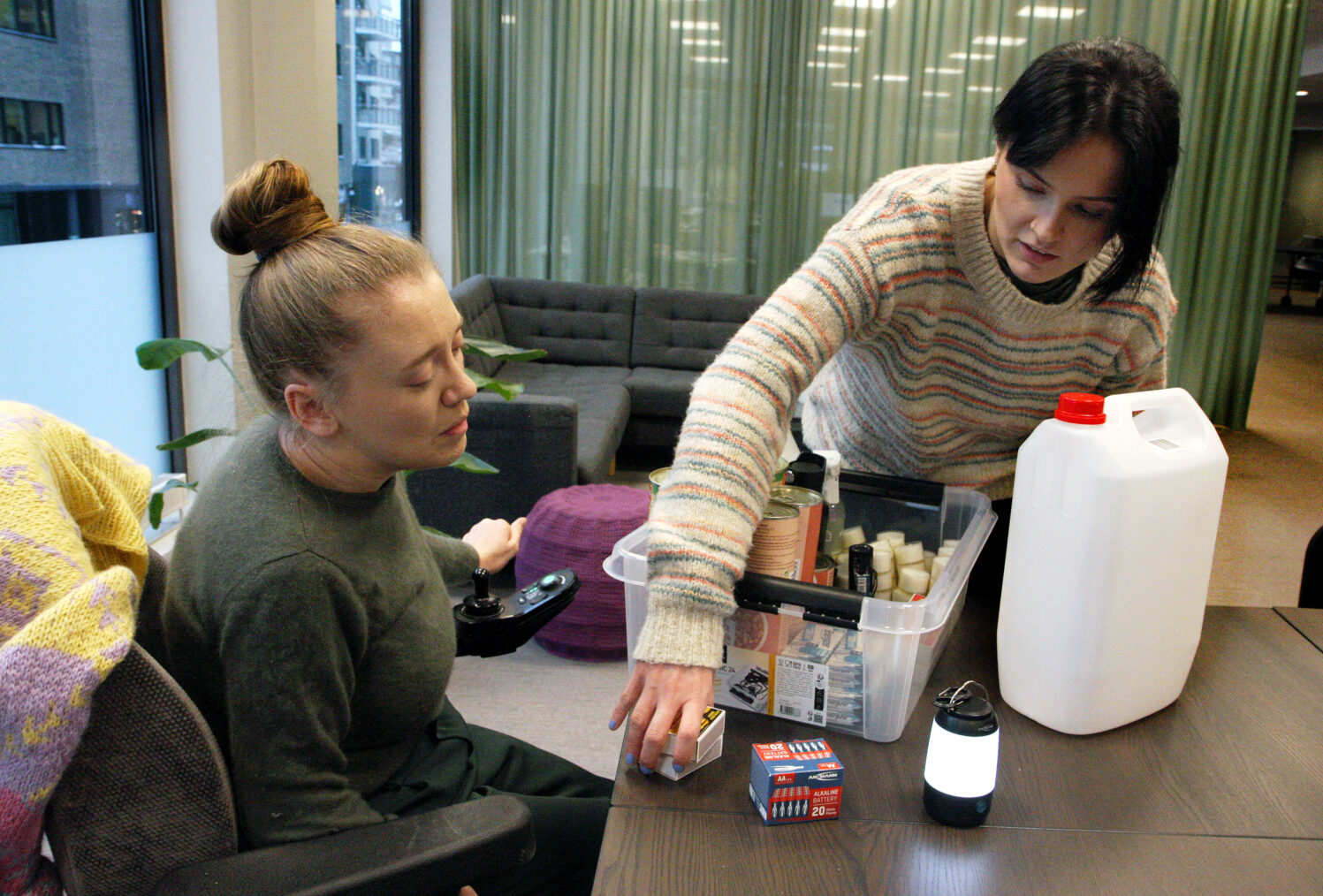 Kvinne i rullestol og assistent fyller plastkasse med varer til beredskapslager