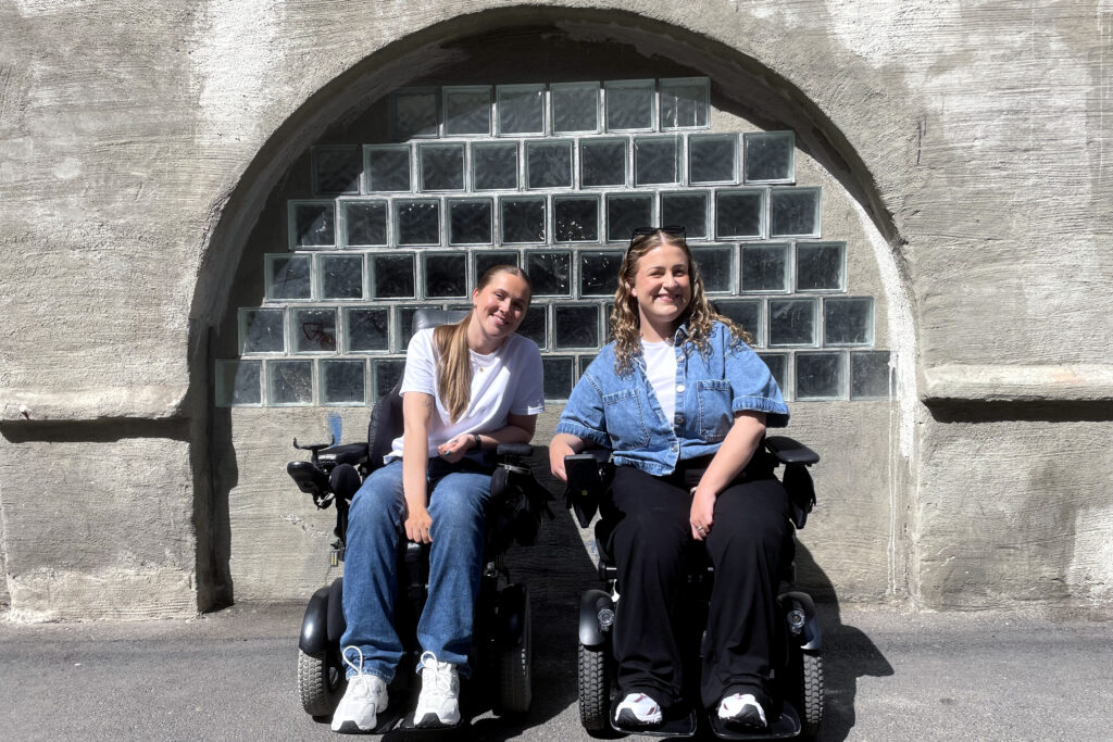 Hannah Lunden og Amalie Rakkestad i rullestoler på fortau foran bue med vindu av glassbyggestein