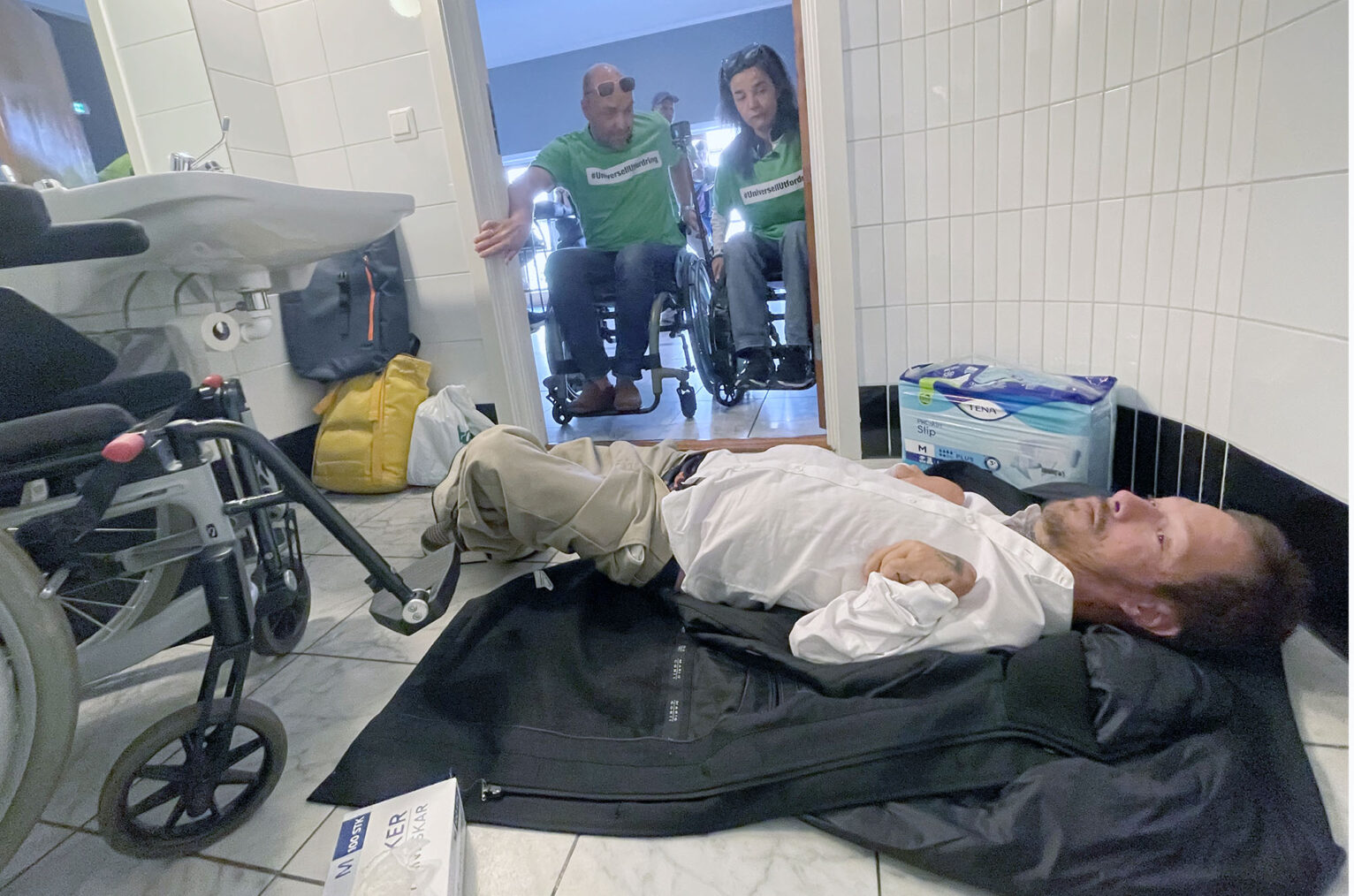 Daniel Blindheim ligger på gulvet på HC-toalett med buksa nesten nedenfor boxershortsen, to personer i rullestol titter inn gjennom døråpningen