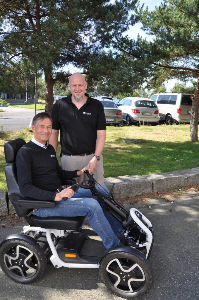 Jan Idar Olsen sittende på scooter foran parkeringsplass, Trone Elvestad står bak