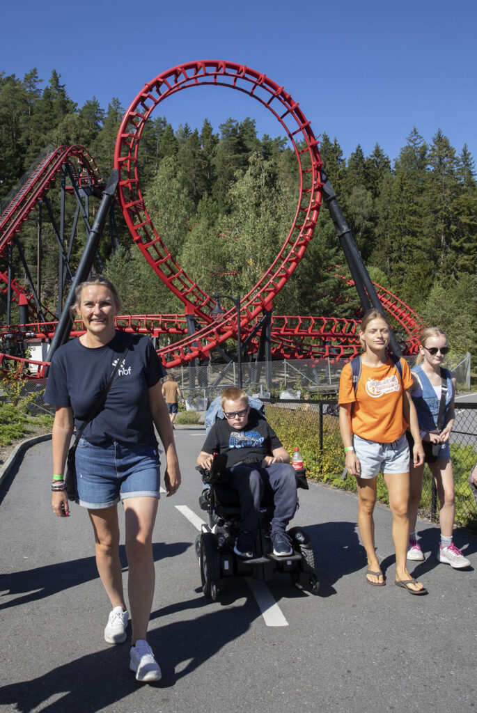 Mor, gutt i rullestol og to jenter smilende på vei bort fra berg- og dalbane