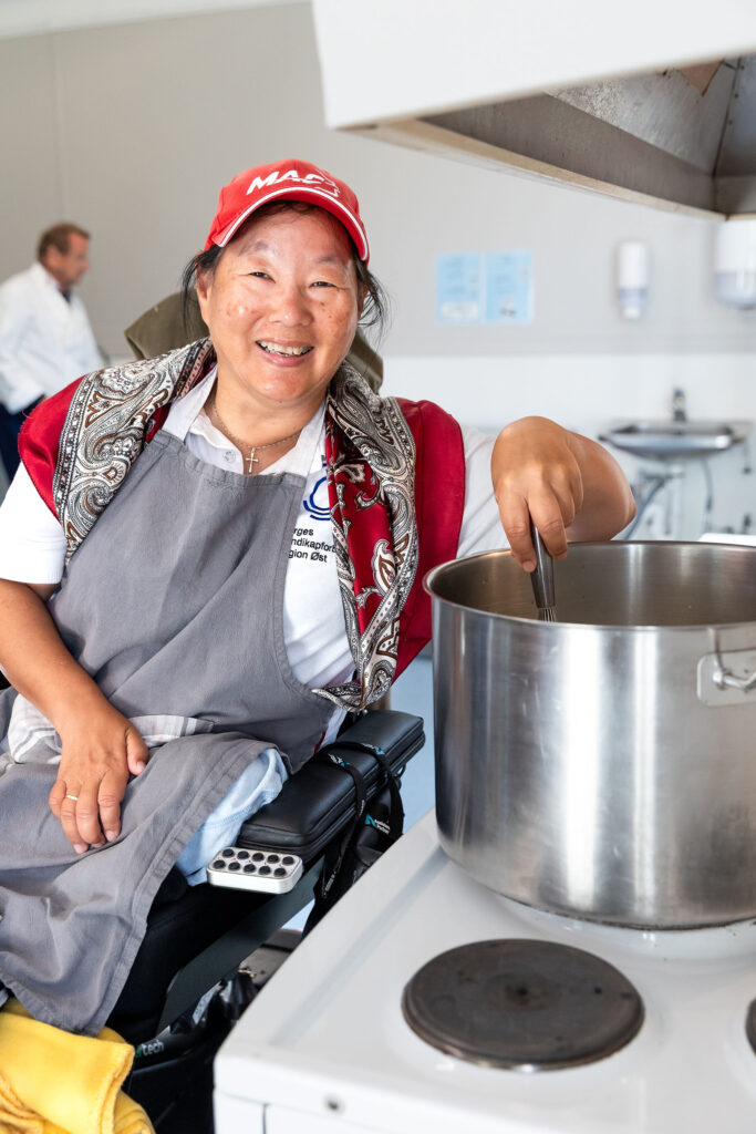 Kvinne i rullestol rører i stor gryte mens hun smiler til fotografen