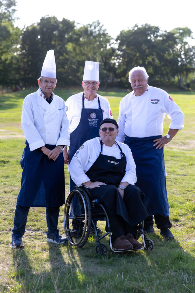Fire menn i kokkeuniform, en av dem i rullestol, på gresslette