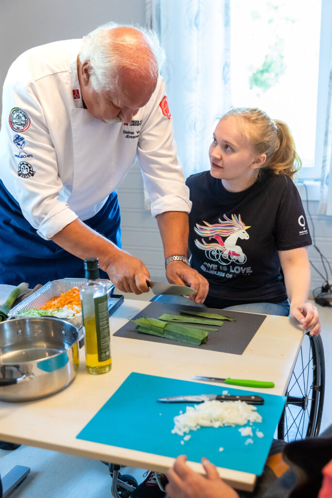 Kokk instruerer ung kvinne i rullestol mens hun kutter grønnsaker