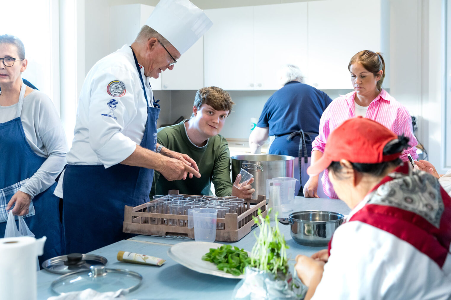 Kokk gir instrukser til tre kursdeltakere på et stort kjøkken.