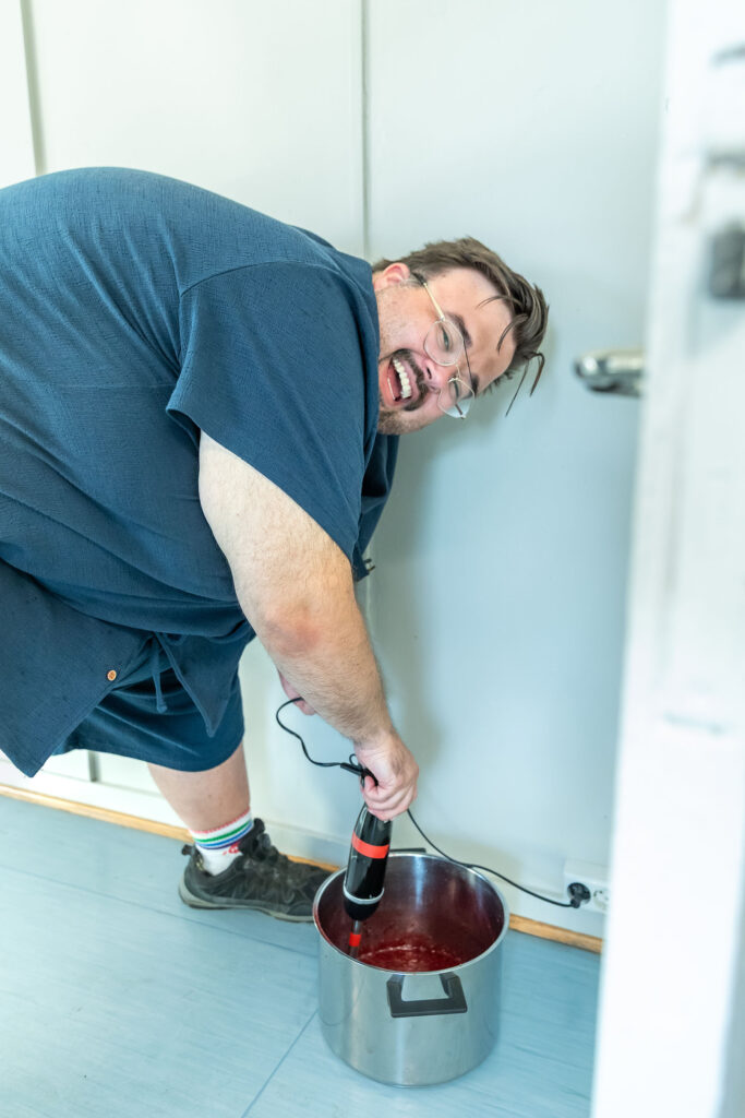 Smilende mann blender saus med stavmikser i en kaserolle som står på gulvet