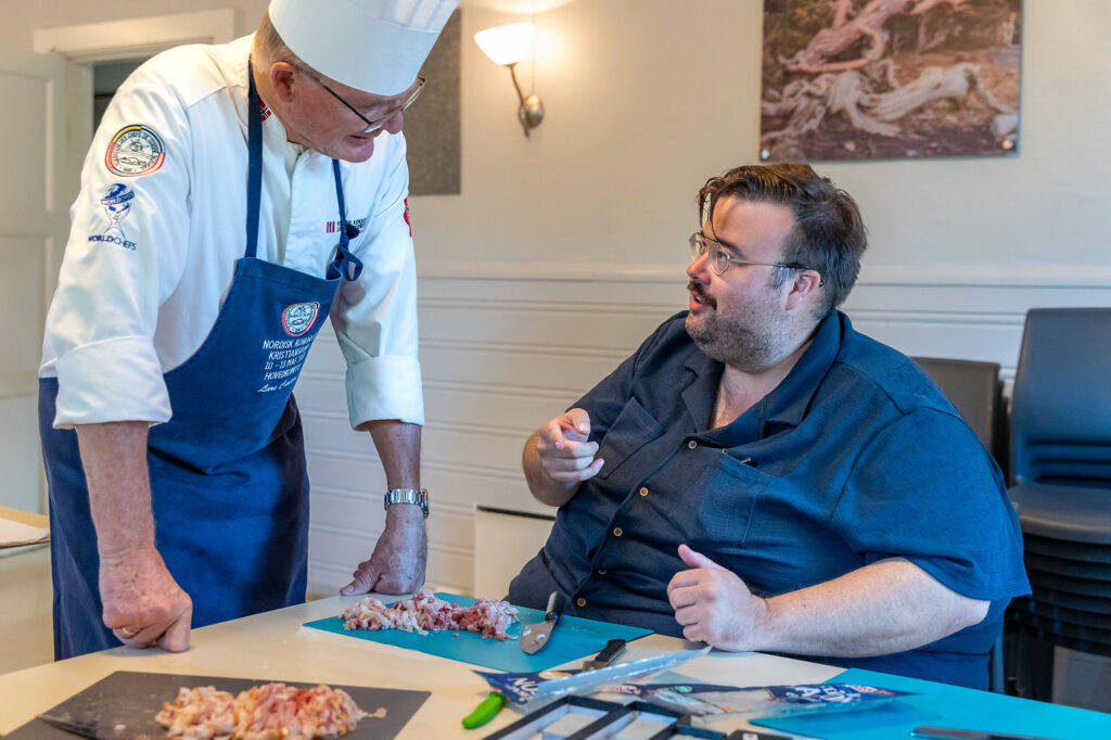 Kokk snakker med mannlig kursdeltaker som kutter bacon