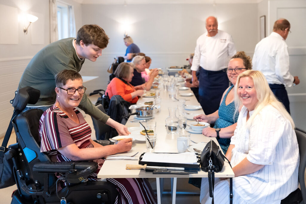 Funksjonshemmede og assistenter ved dekket langbord, kokker går i bakgrunnen