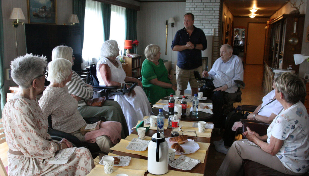 Øyvind Ekvik stående ved salongbord med mange eldre mennesker rundt