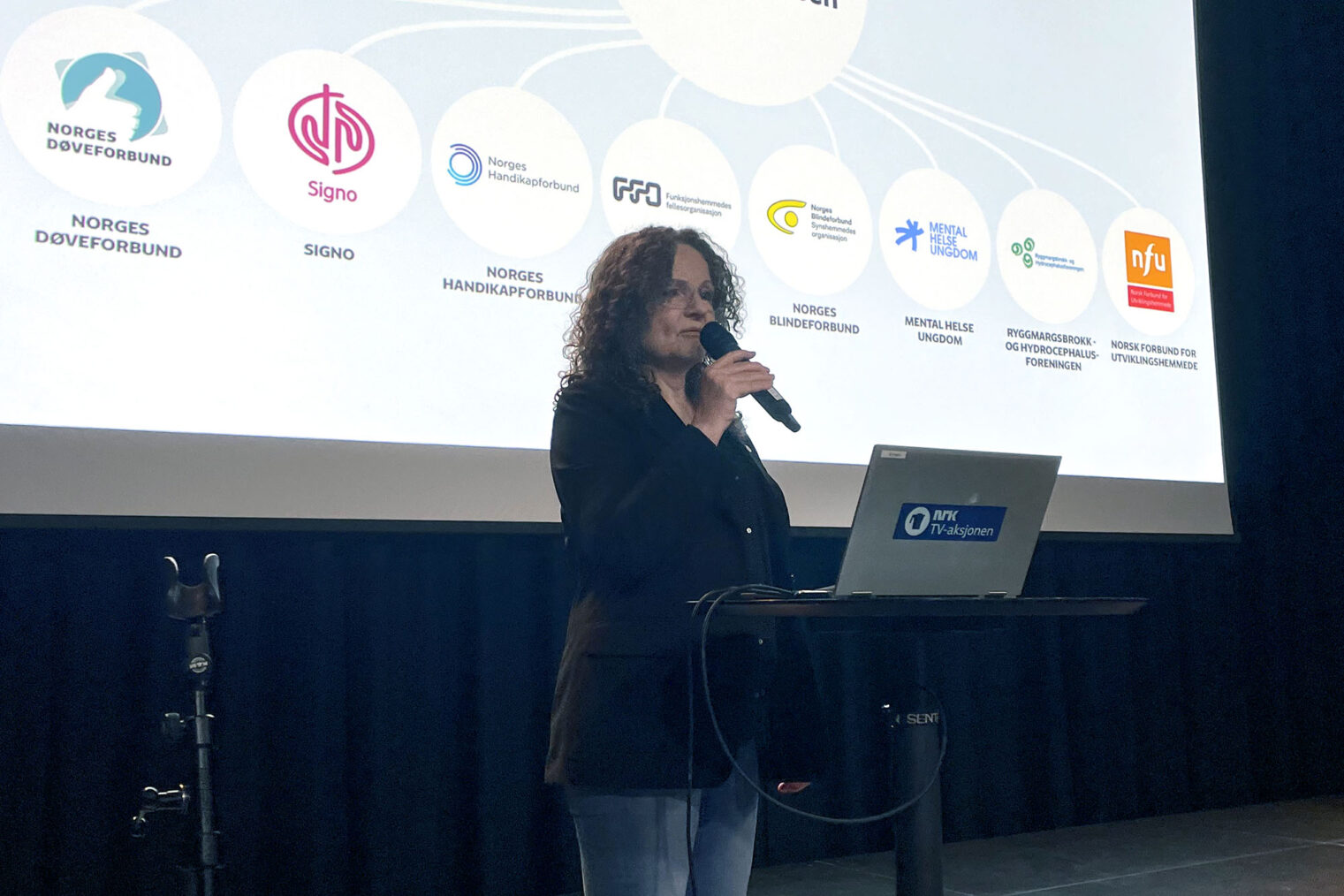 Marit Sørheim på scene foran storskjerm som viser logoen til sju organisasjoner i Atlas-alliansen