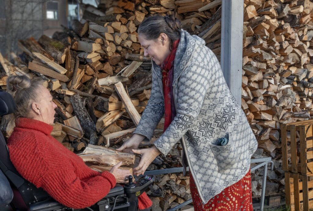 Foren store vedstabler legger Frøydis Stiansen vedkubber i fanget til Maria Hagen