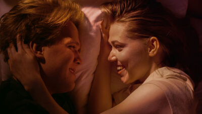 Ung mann og kvinne ligger i dobbeltseng og smiler til hverandre, kvinnen stryker nakken til mannen