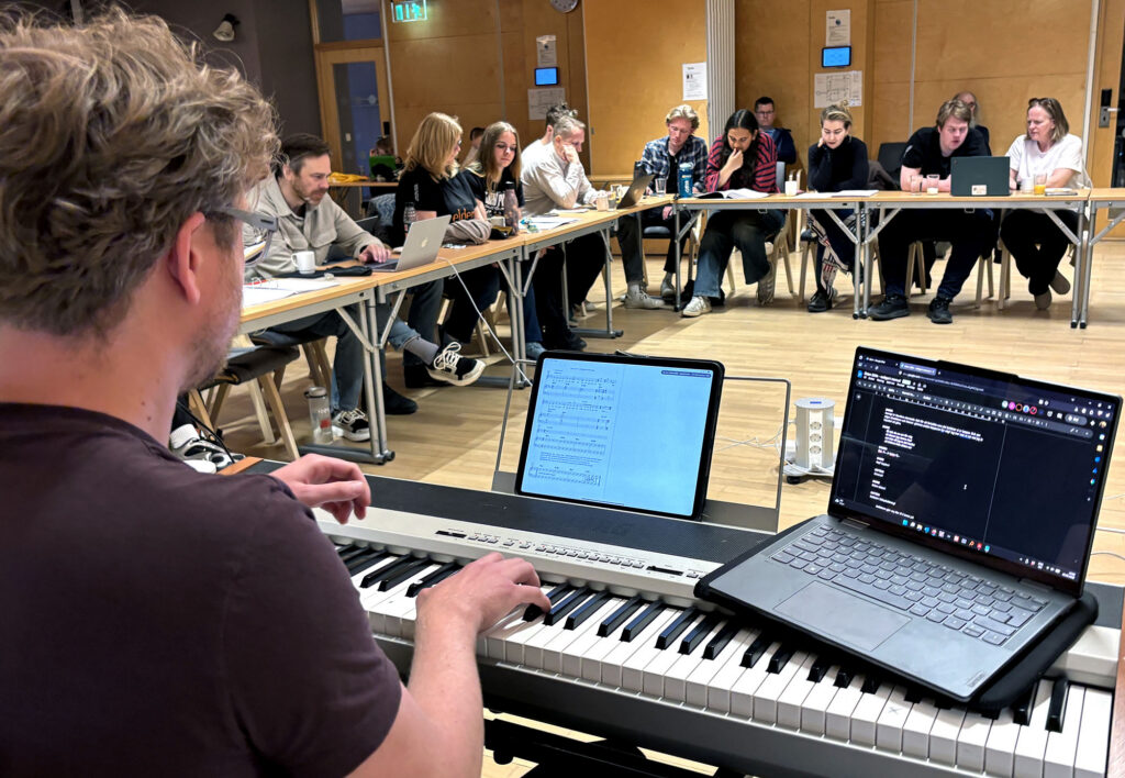 Mann med keyboard og to laptoper foran skuespillerteam sittende ved bord i hesteskoform i seminarrom