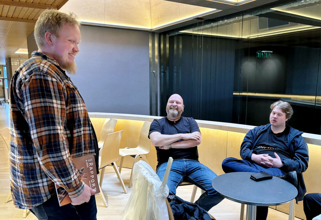 Stian Tranung står og snakker med Jo Inge Nes og Christoffer Bratseth, som sitter ved et bord.