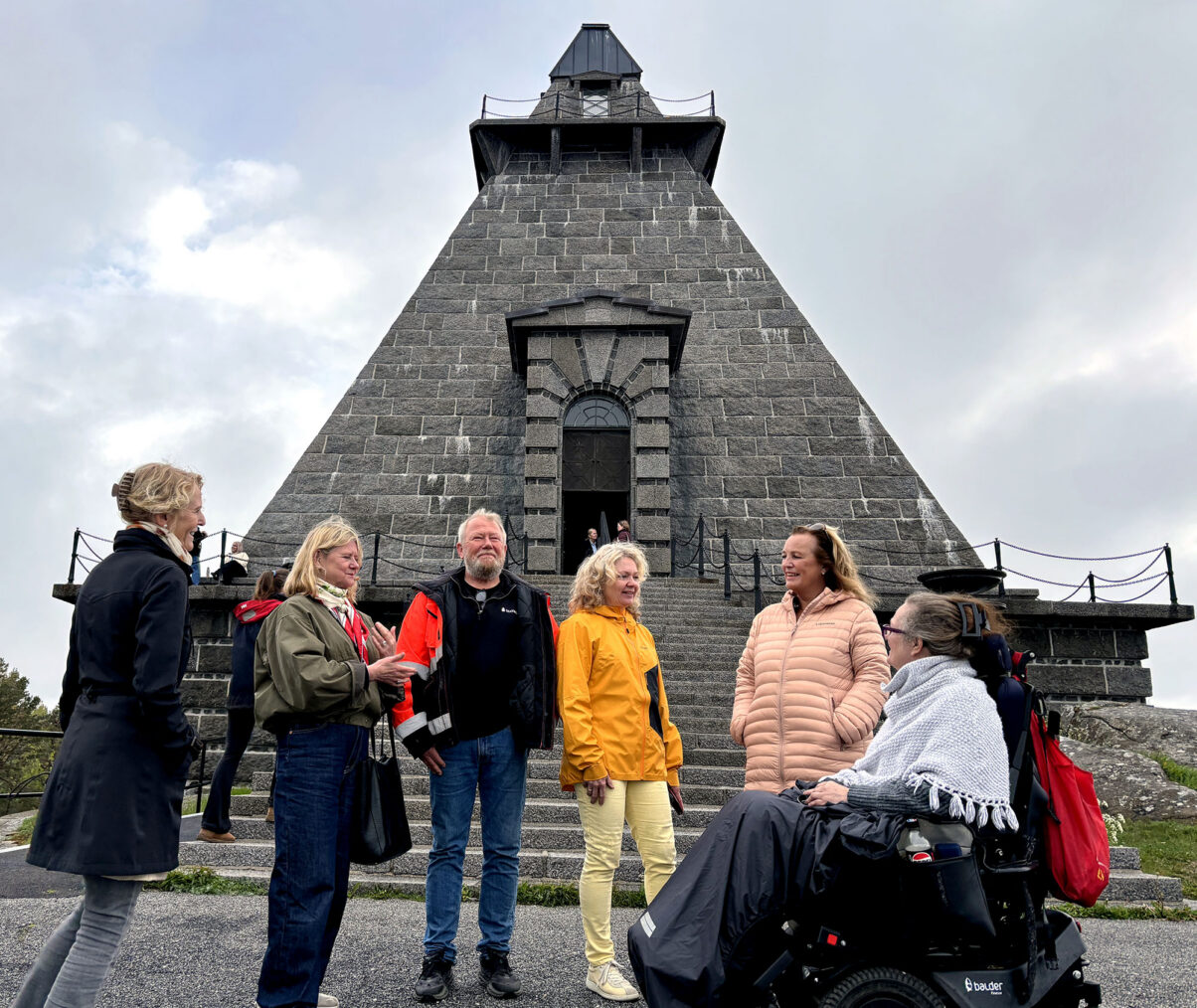 Seks mennesker, hvorav en kvinne i rullestol, foran den pyramideformede steinbygningen Minnehallen