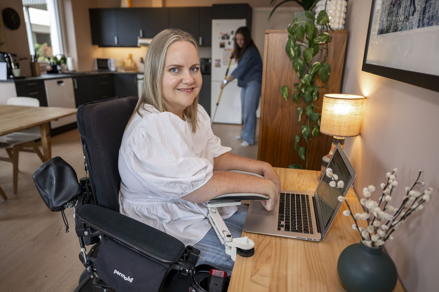 Anne Torunn Sirevåg skriver på PC mens assistent vasker gulvet på kjøkkenet i bakgrunnen.