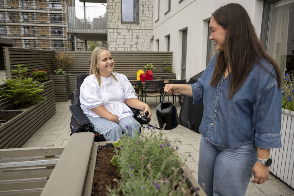 Assistent vanner blomster i terrassekasser, Anne Torun sitter i bakgrunnen, de smiler til hverandre.