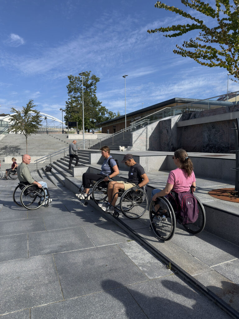 Fem personer i manuell rullestol på hellelagt plass snakker sammen